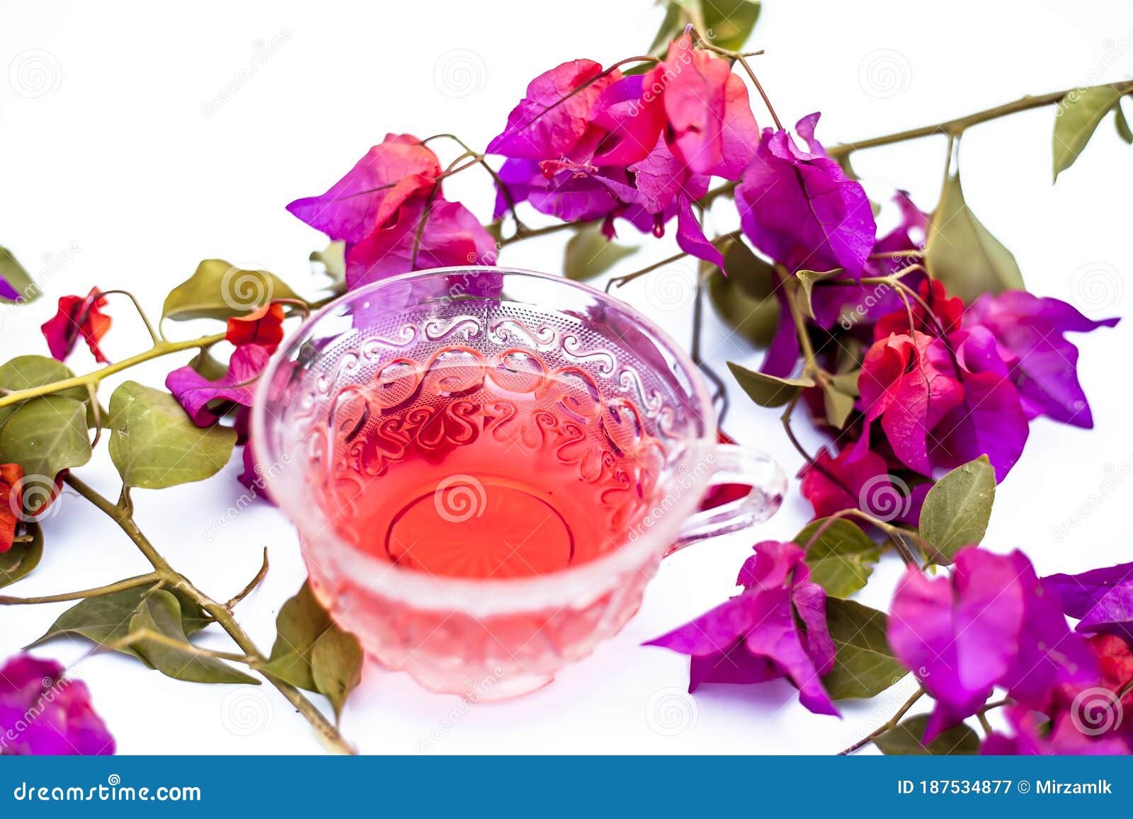 Secangkir seduhan teh herbal bening berwarna kemerahan dari kelopak bougenville kering bersanding dengan teko kaca dan piring kayu kecil berisi bunga kering.