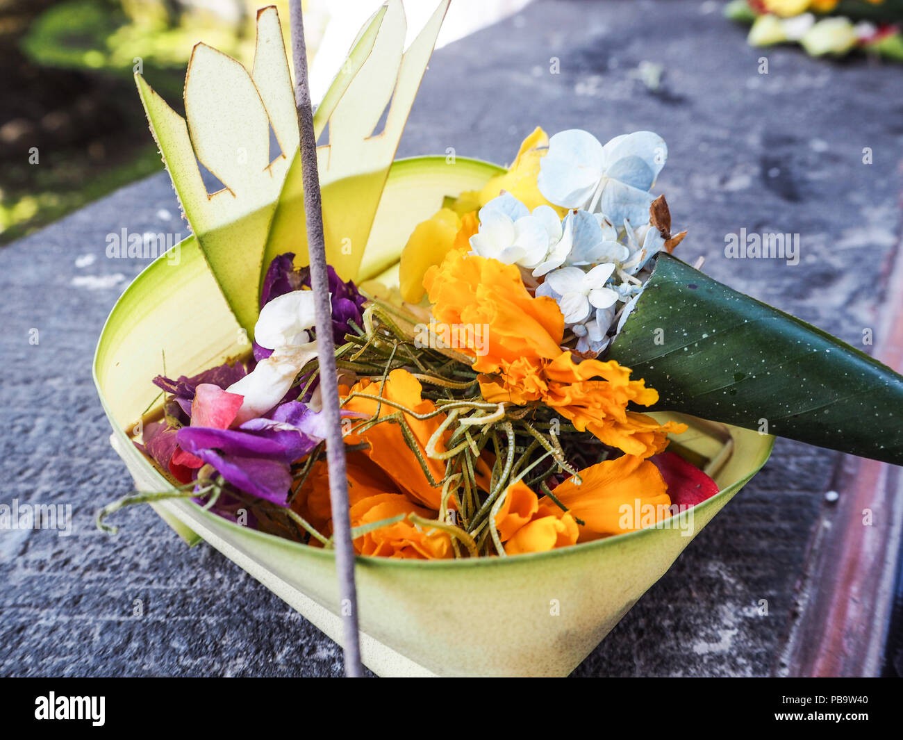 Anyaman daun kelapa (canang sari) berisi tumpukan kembang asoka jingga cerah, kamboja, dan taburan irisan daun pandan wangi di atas pelataran pura.
